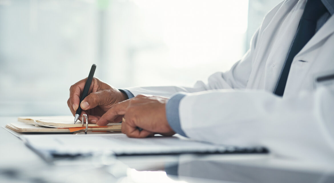 Doctor hands writing on paper or document at a desk in the hospital. Healthcare professional drafting a medical insurance letter, legal paperwork or form. A GP filing a document in a clinic office
