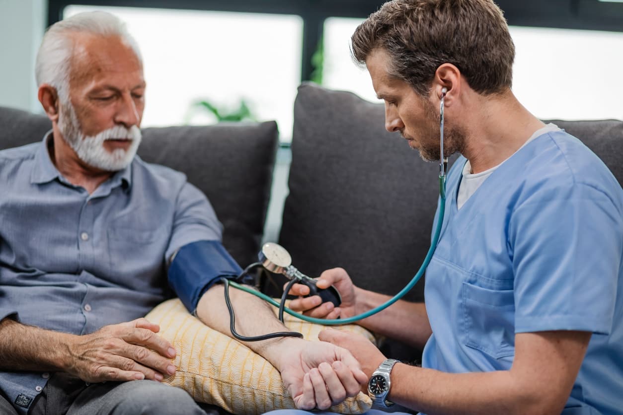 Male caregiver checking blood pressure of senior man at home with sphygmomanometer during healthcare visit