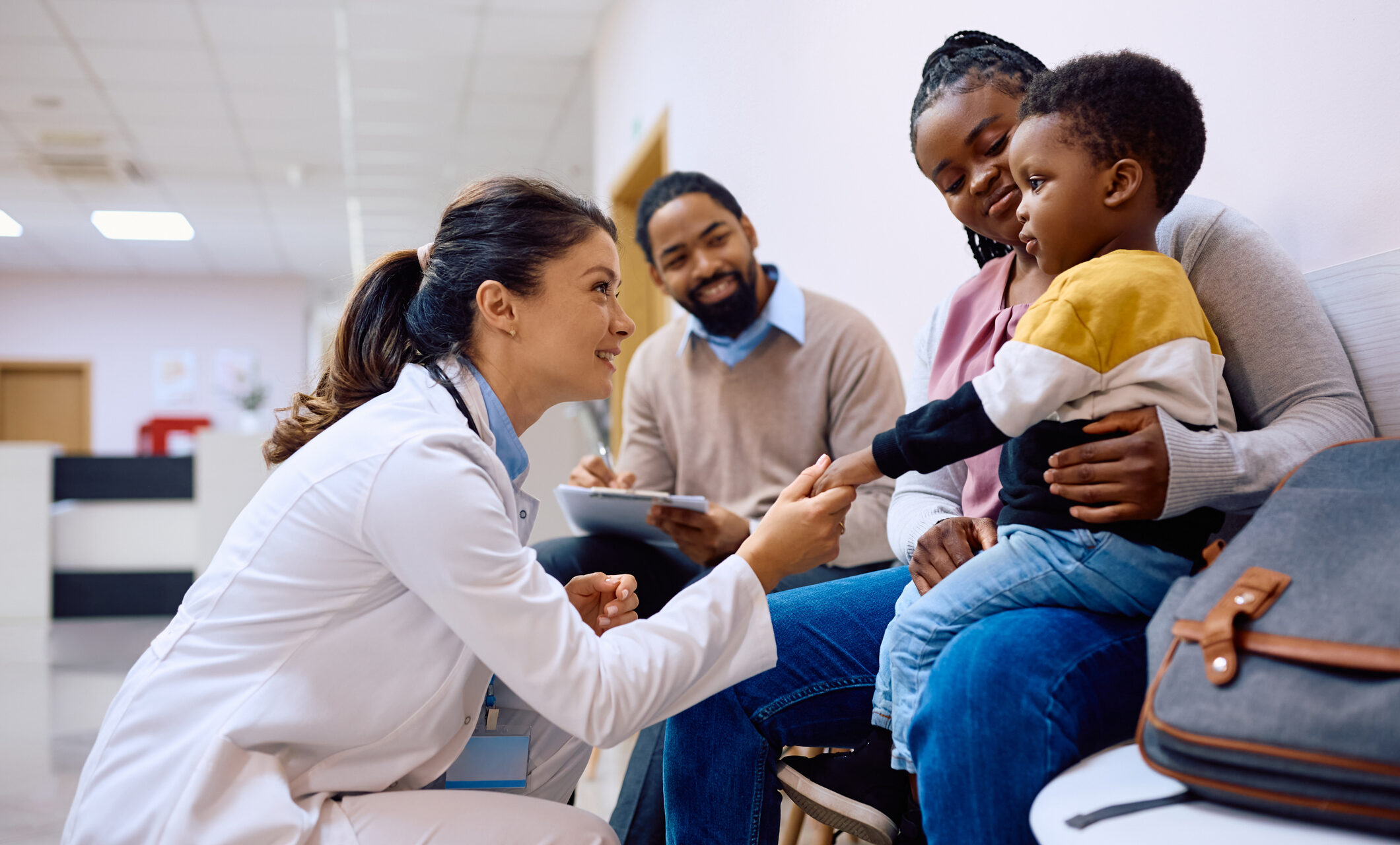 Smiling doctor holding hand on African American boy who came with parents for medical exam at pediatric clinic.