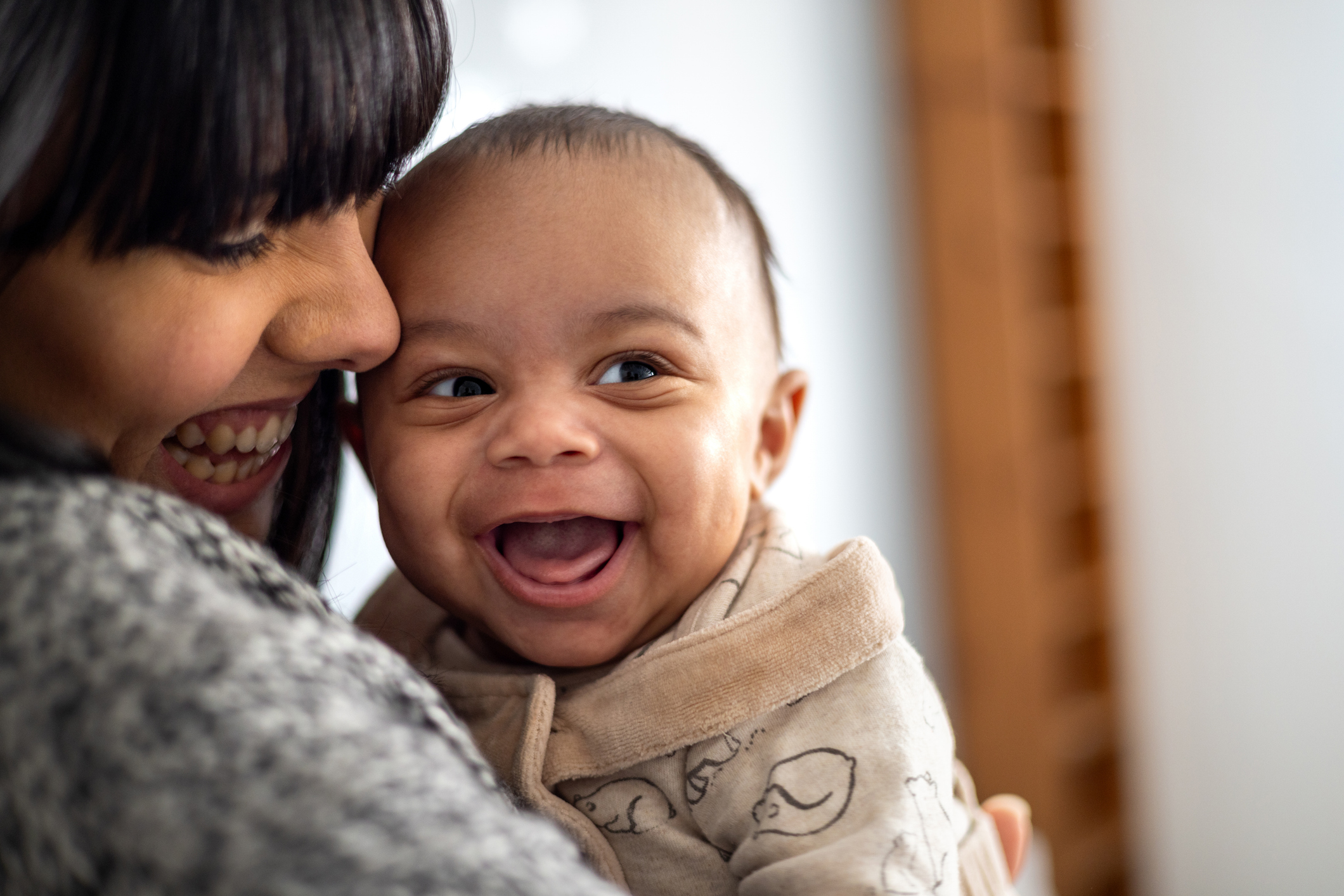 Close-up of a mother holding her baby boy near her face. The baby boy is laughing looking away from the camera making his mother laugh.