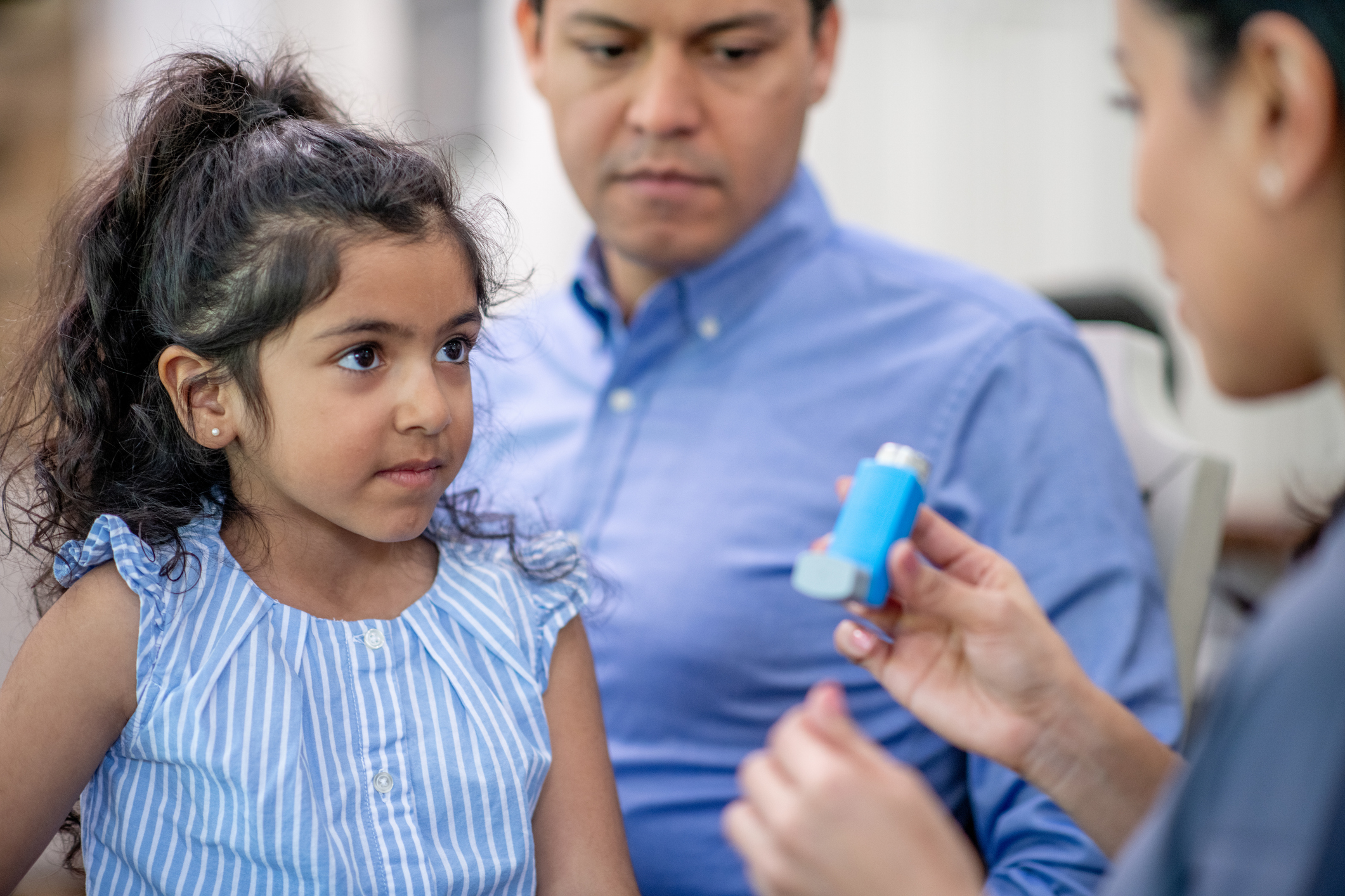 A Father brings his daughter in to the doctor to address his concerns with her Asthma. The female doctor is holding out an inhaler as she reviews the proper administration with the the young girl.