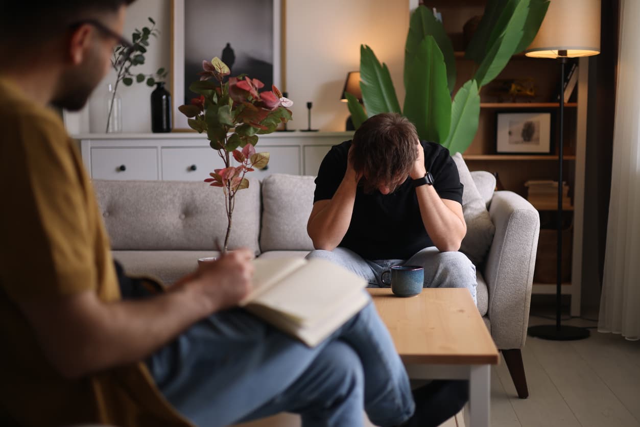 Man sitting on a couch during a mental health counseling session with a therapist taking notes in a modern office.