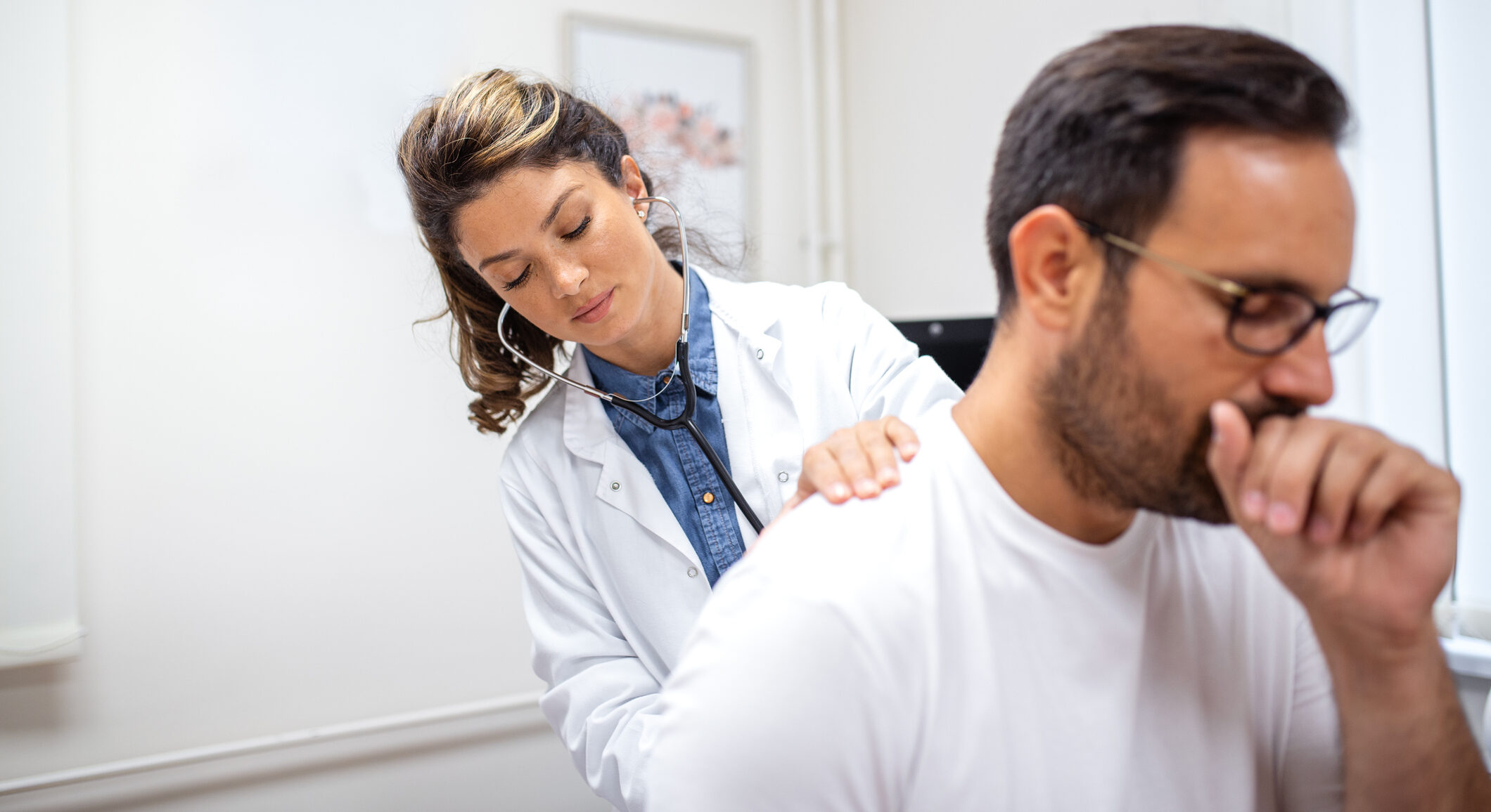 Female doctor listens to male patient's back with a stethoscope as he coughs. The doctor is focused while the patient shows visible discomfort, suggesting respiratory symptoms.