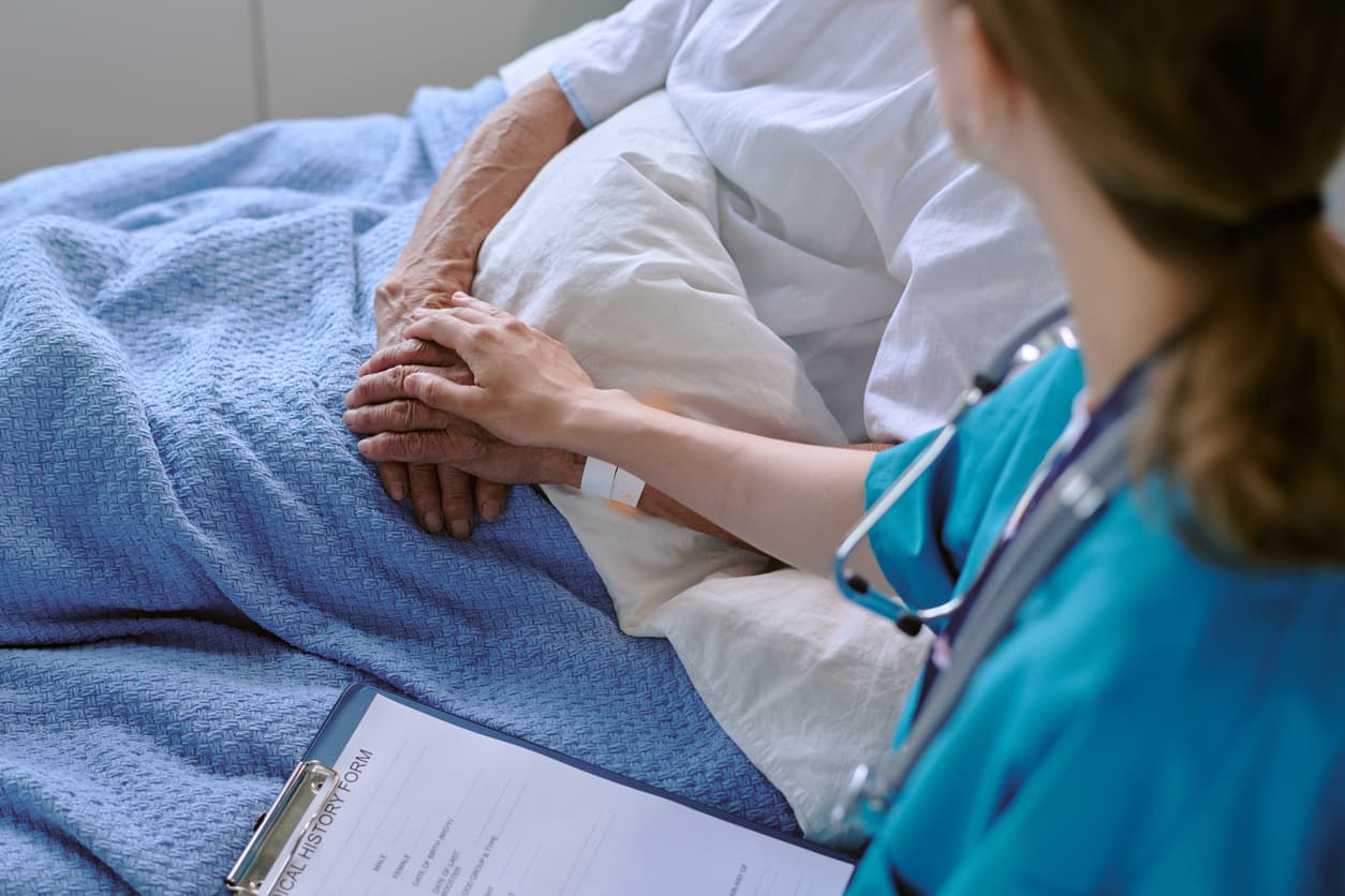 Hospice nurse comforting elderly patient in bed by holding hand during compassionate end-of-life care visit