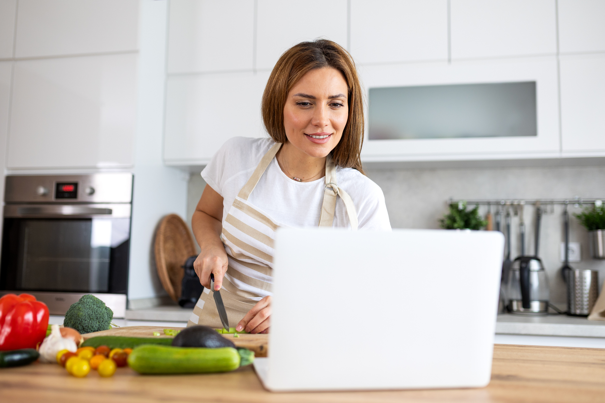 Online cooking class. Woman following recipe on laptop, Digital technology in kitchen, Education and hobby concept. Attractive lady in apron cutting vegetables while looking at computer screen.