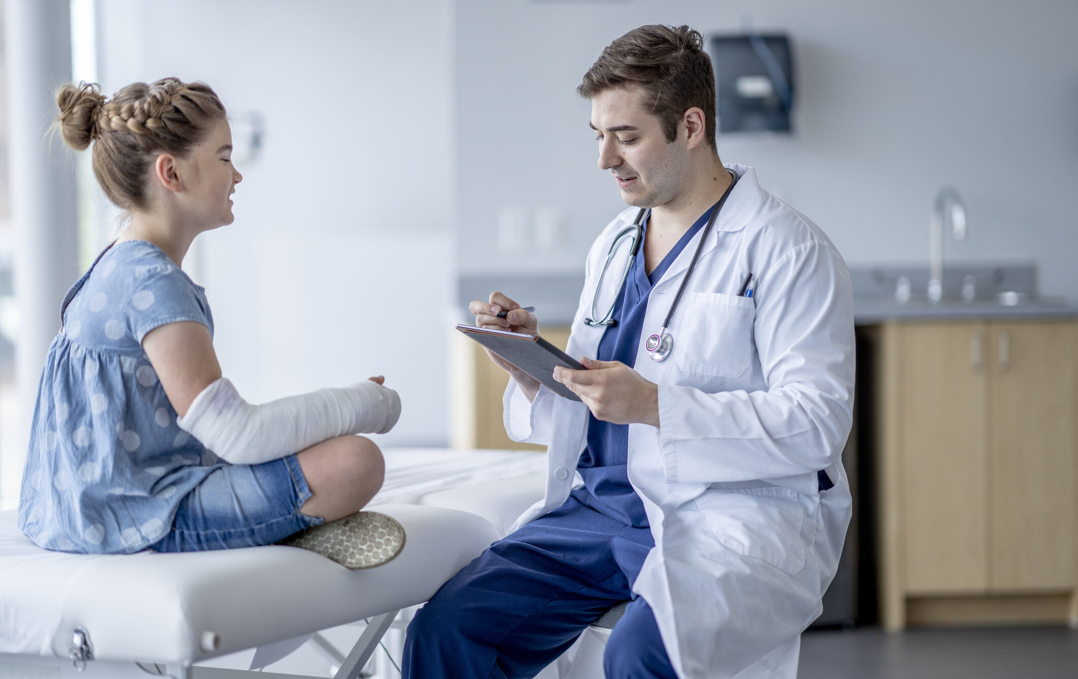 A sweet little girl with a broken arm, sits up on an exam table as she talks with her Orthopedic surgeon about her arm.