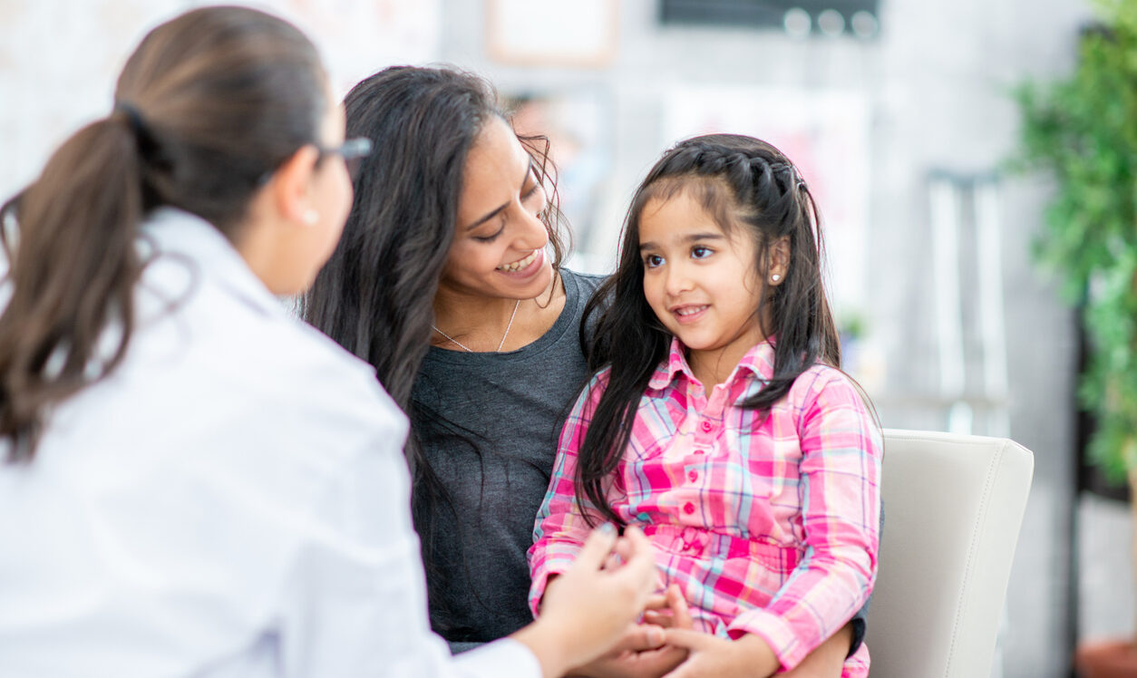 A Mother and her Daughter sit in a medical office across from their Pediatrician during an appointment.  The little girl is sitting on her Mothers lap and is smiling as they talk with the doctor.