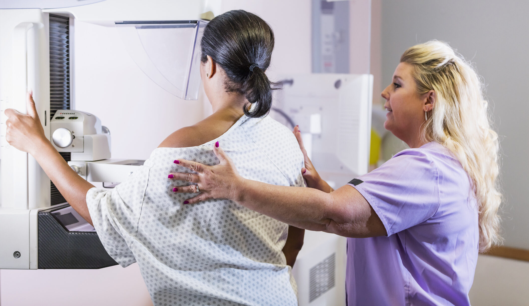 A mature African-American woman in her 40s wearing a hospital gown, getting her annual mammogram.  She is being helped by a technologist, a blond woman wearing scrubs.
