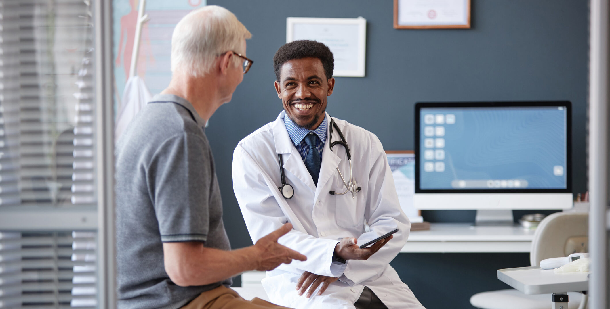Smiling Black Doctor Holding Tablet Talking to Senior Patient in Consultation