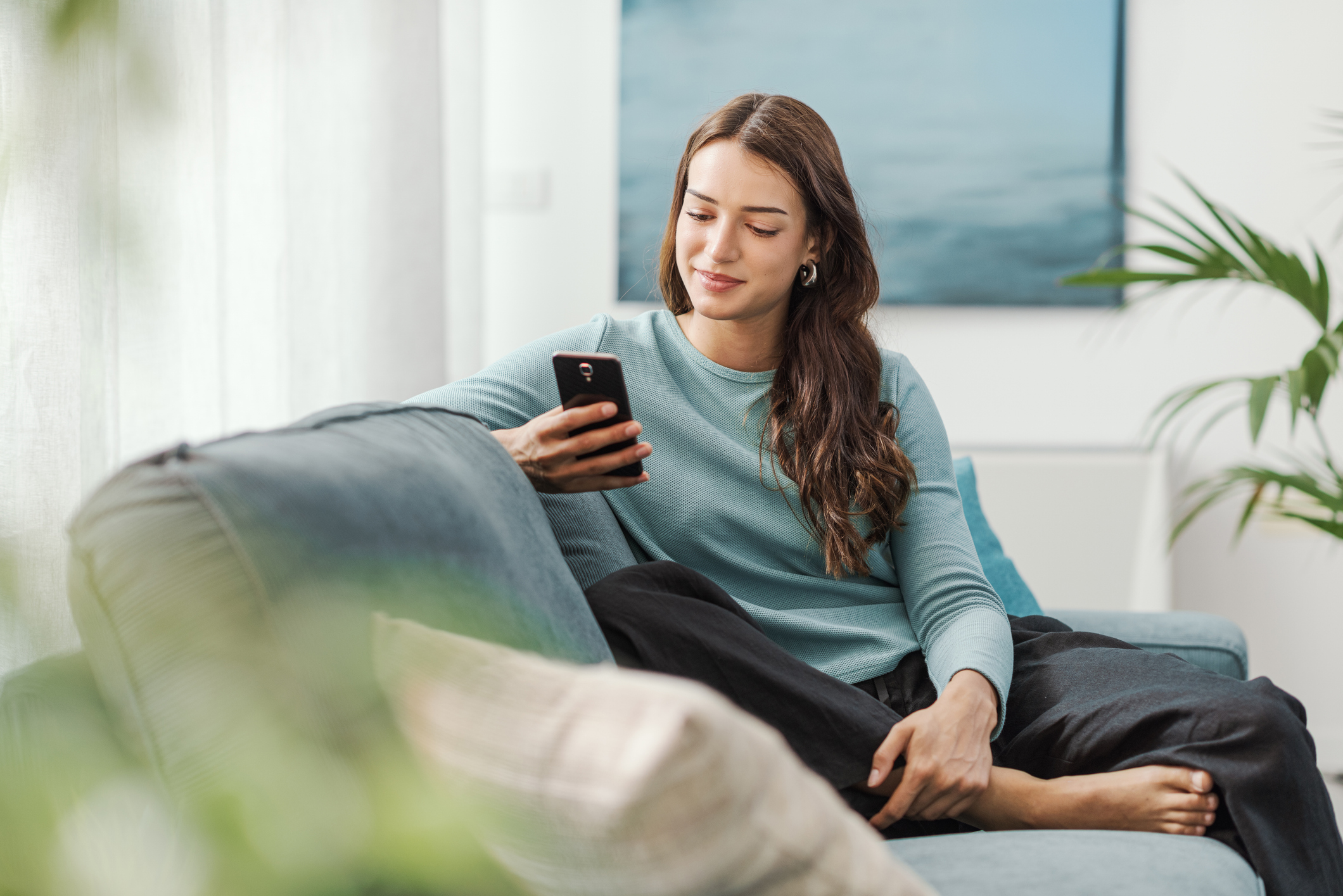 Happy young woman sitting on the couch at home and chatting with her smartphone, she is social networking and using mobile apps