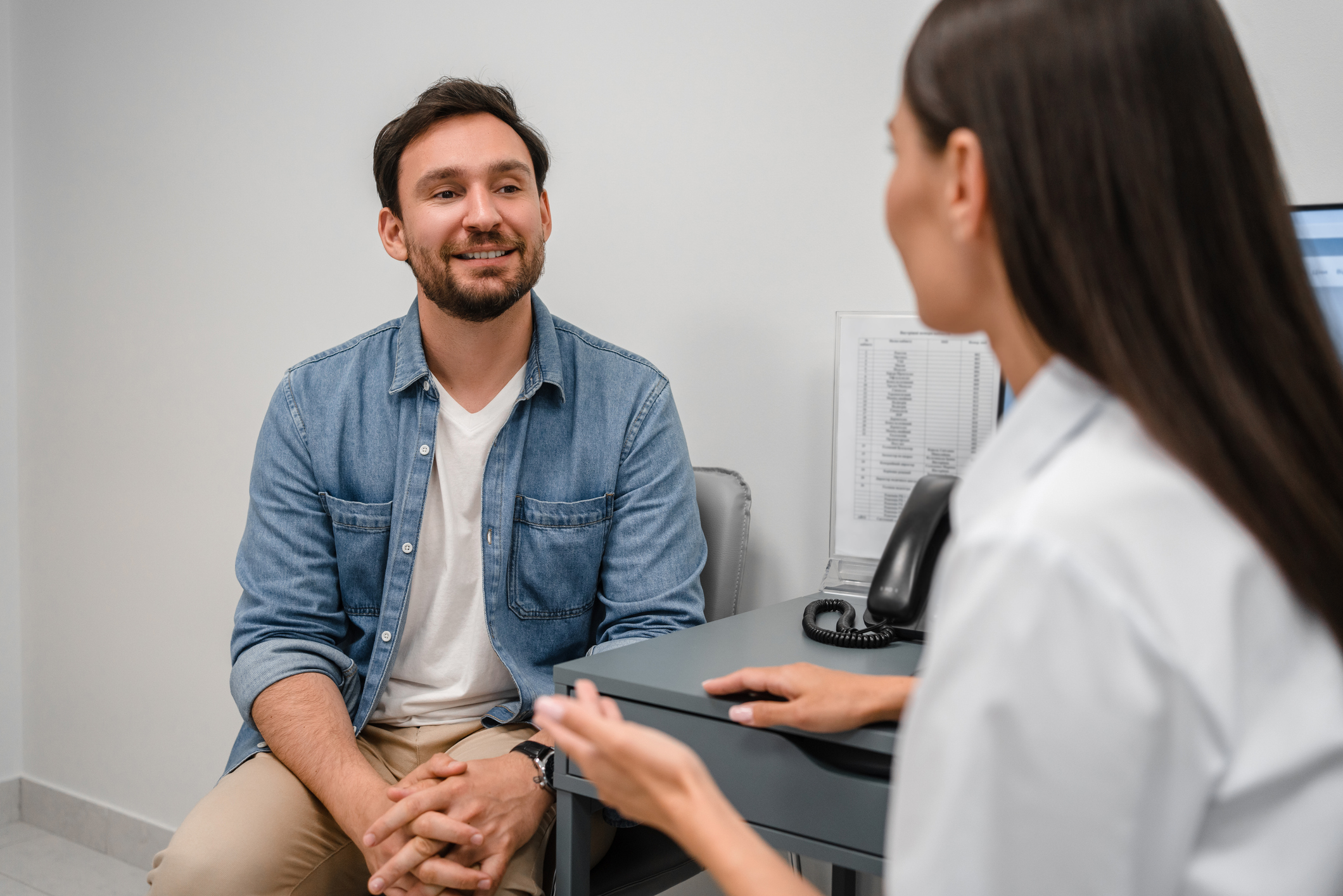Smiling woman doctor listening to male patient complaints at meeting in hospital. Friendly female therapist physician wearing white uniform consulting young man at medical appointment