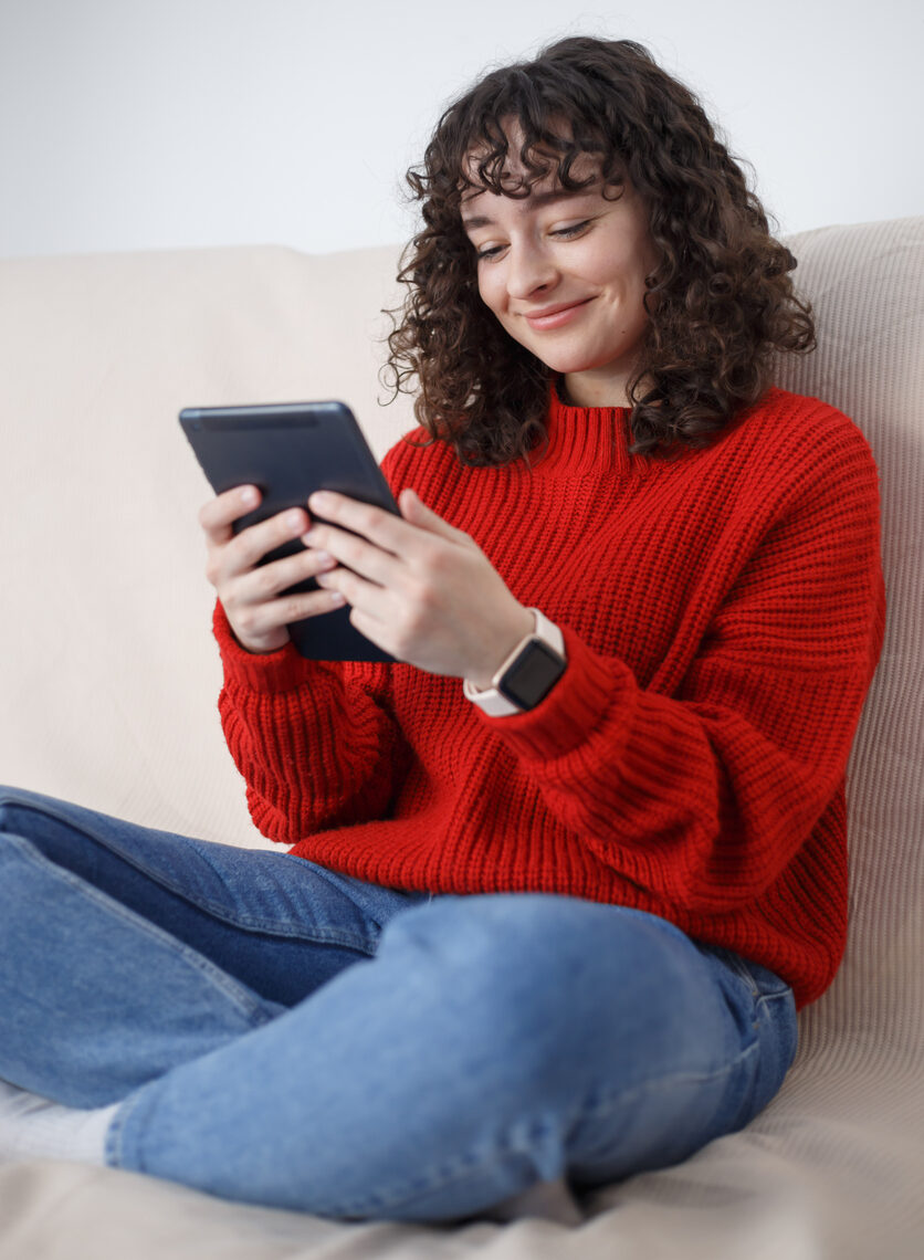 Cheerful curly female person sitting on sofa at home and browsing internet on portable gadget