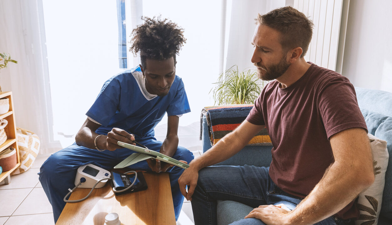 A healthcare worker in blue scrubs sitting on an orange stool, attentively using a digital tablet during a medical consultation with a patient. The patient, dressed in a maroon shirt and blue jeans, is seated beside him on a blue sofa that features a white cushion and a colorful striped throw. They are in a bright, welcoming living room with white sheer curtains diffusing sunlight across the room. A blood pressure monitor, a bottle of medication, and a notepad rest on a light wood coffee table. In the background, there is a comfortable home environment with a shelving unit containing plants and knick-knacks, reflecting a personal.