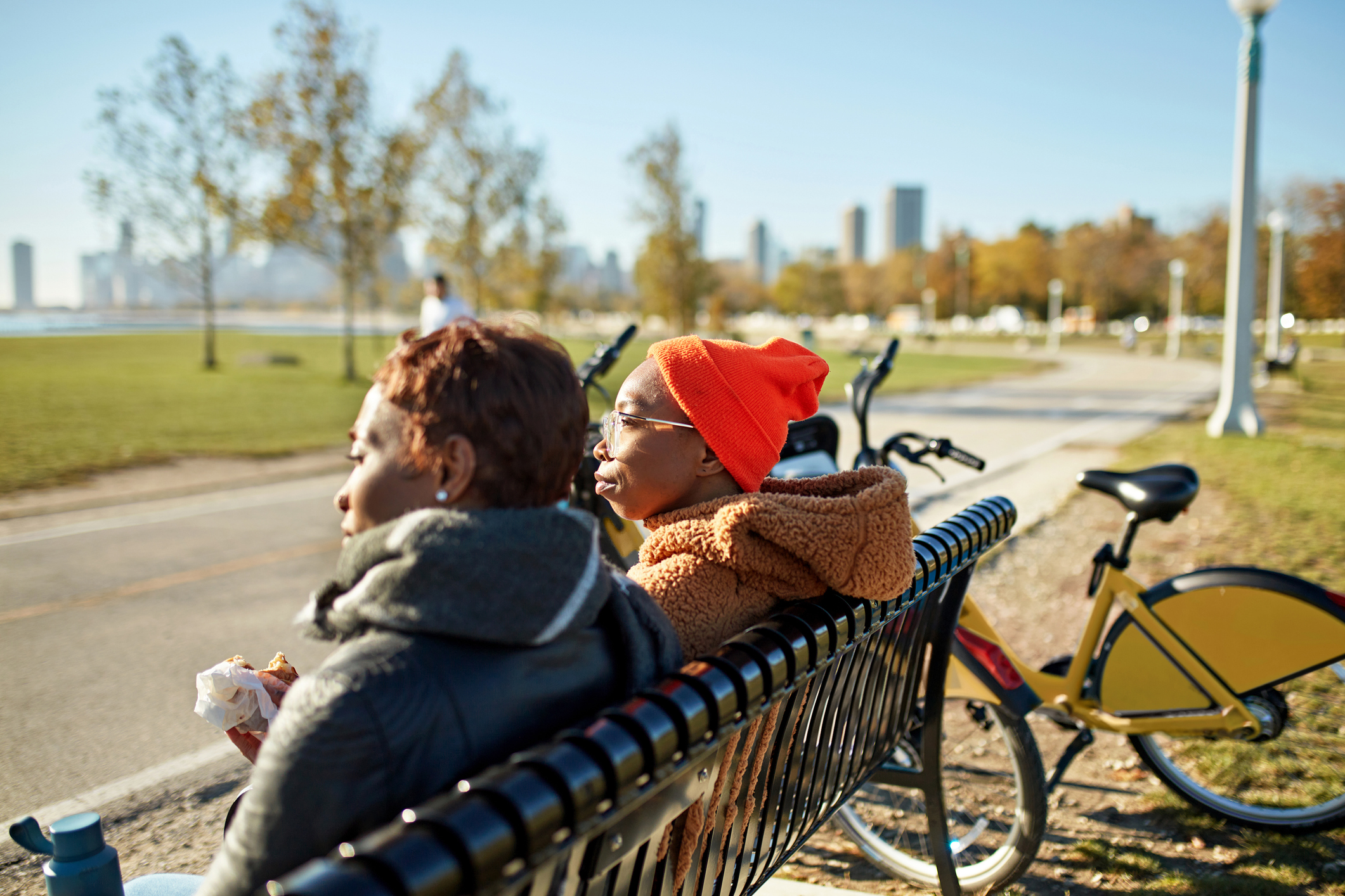 Chicagoans in 20s and 30s wearing warm clothing, sitting side by side on park bench, taking a break from bike riding for snack and view of lake.
