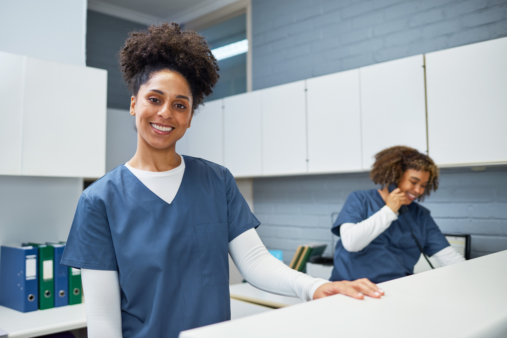A cheerful and friendly healthcare worker wearing a uniform is standing at a reception desk in a medical facility, smiling warmly. She is supported by a colleague working in the background.